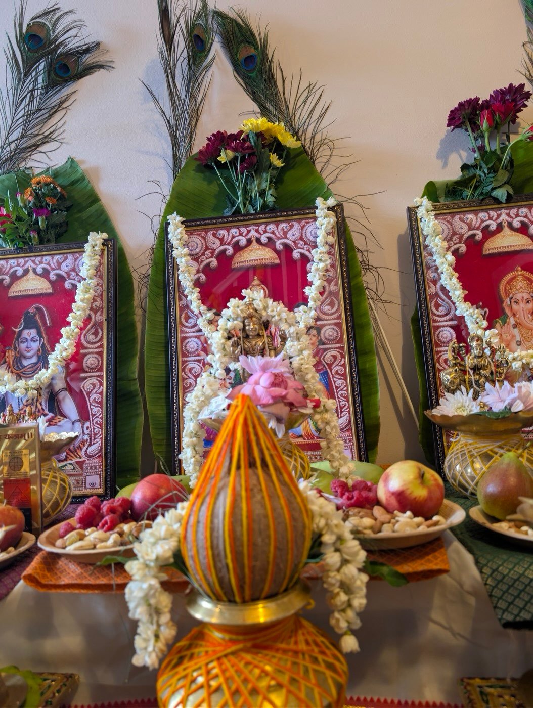 Kalash wrapped in orange thread in front of framed deities and banana leaves